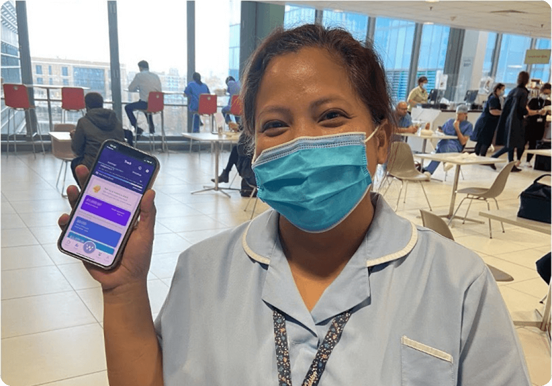 Smiling healthcare worker holding a smartphone displaying the Wagestream app, showcasing its financial wellbeing features in a hospital staff room.
