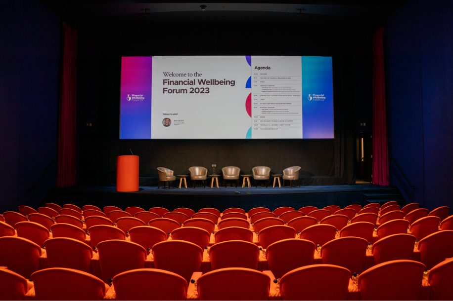 Financial Wellbeing Forum 2023 conference room with orange seating, stage, and presentation screen displaying the event agenda.
