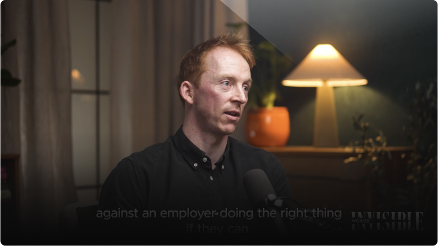 Man in a dark shirt speaks during an interview, a lamp is visible in the background.
