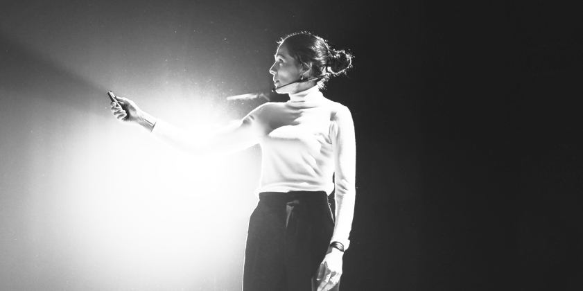 Woman giving a presentation, holding a clicker, illuminated by a spotlight.
