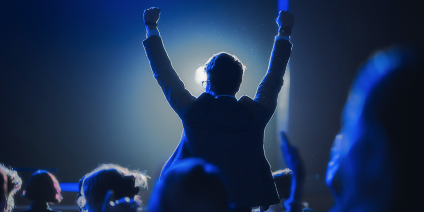A person with arms raised in celebration at a corporate event, backlit by a bright light.
