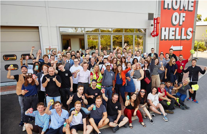 Large group of happy Beer52 employees celebrating outside their brewery, holding cans of beer.
