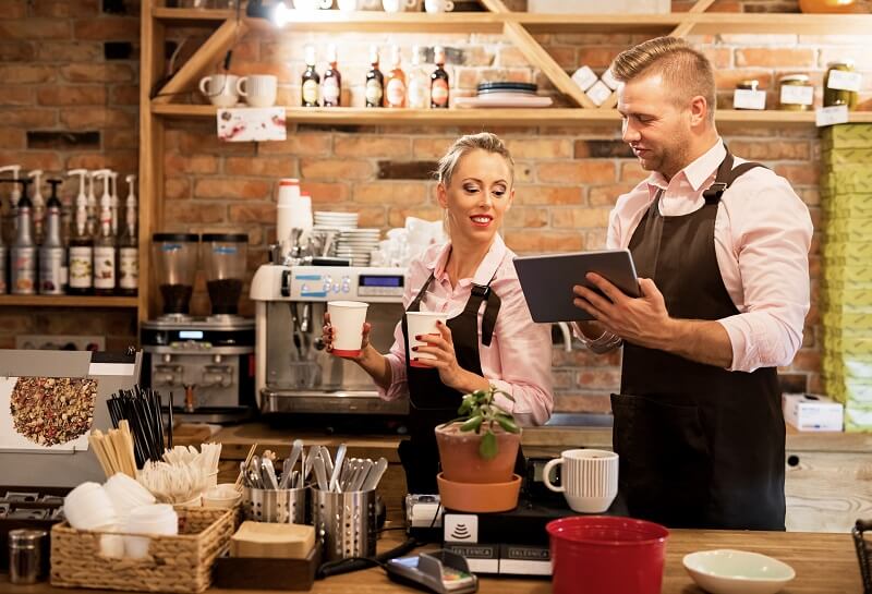 Two baristas working in a cafe, one holding two takeaway coffee cups and the other using a tablet.
