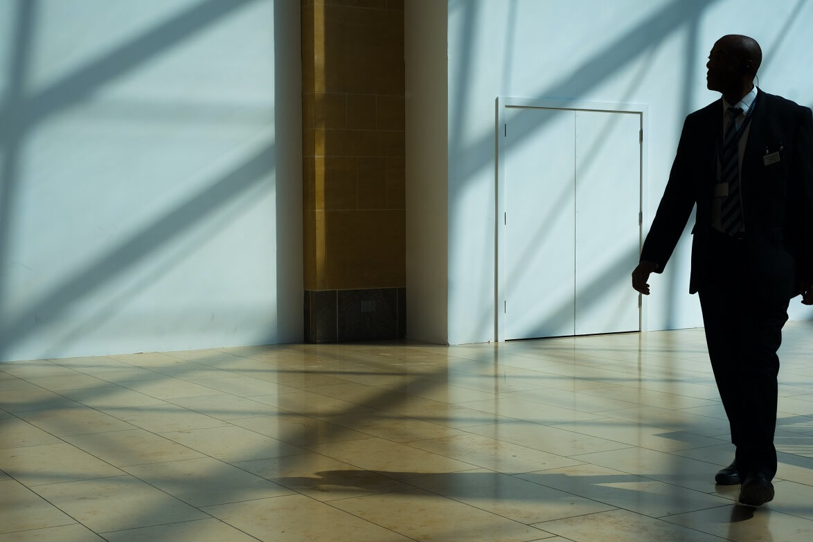 Businessman walking through modern building, sunlight casting shadows on floor.
