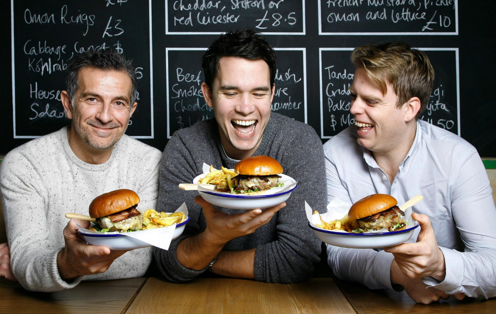 Three men smiling and holding plates of burgers and fries.
