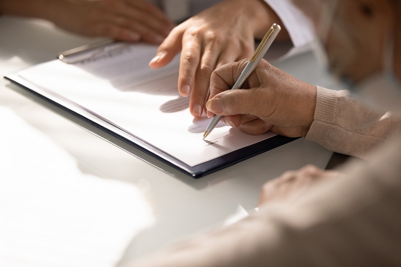 Close-up of a person signing a document with a pen, assisted by another person.
