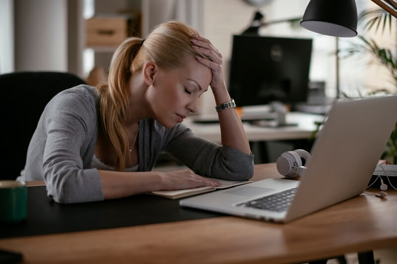 Tired woman leaning on desk, hand on forehead, looking at notebook, laptop in front of her.
