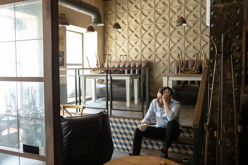 A man sits on steps in a cafe, holding a coffee cup, looking stressed.
