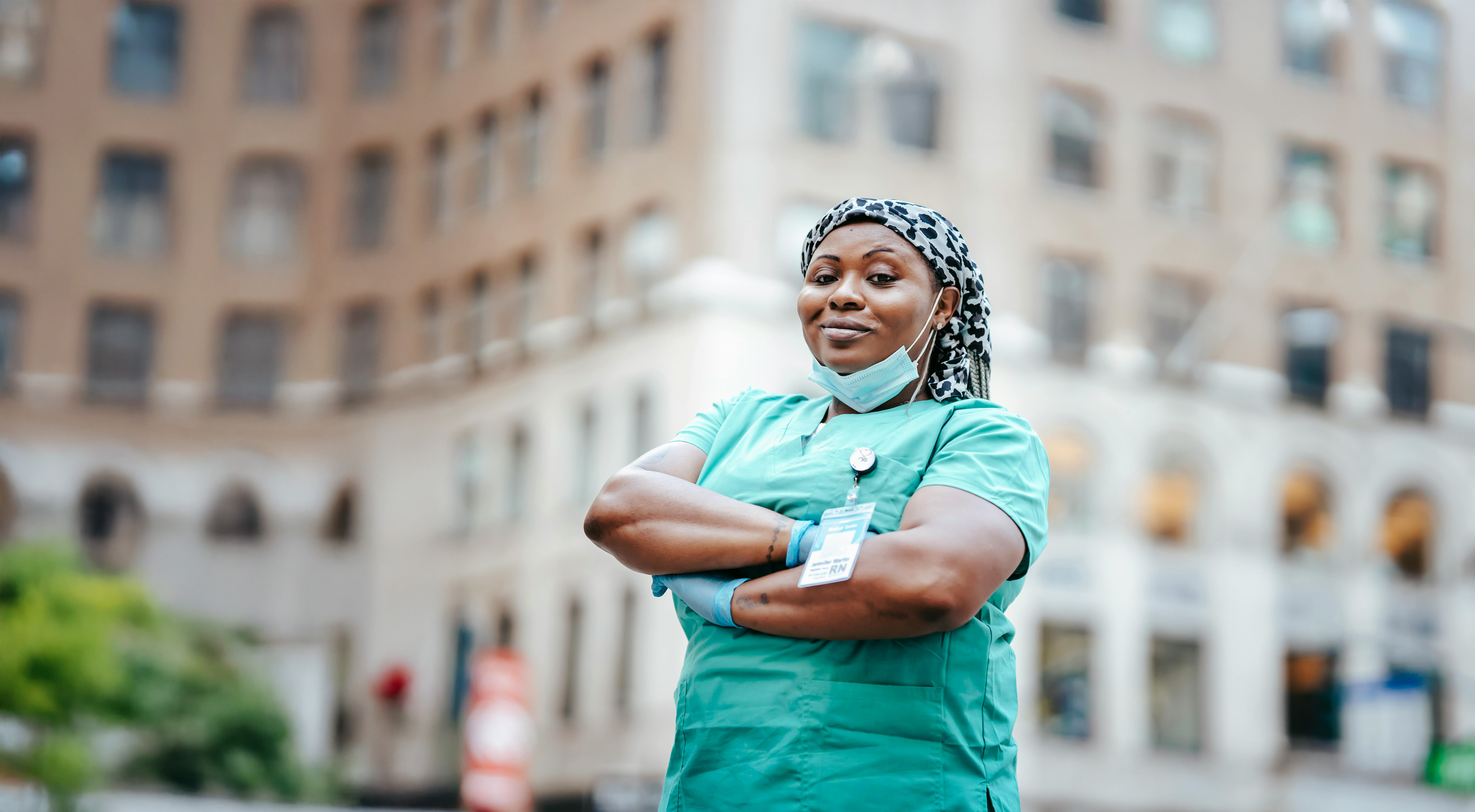 Confident nurse wearing green scrubs and a patterned headscarf standing with arms crossed outdoors in front of a city hospital building.