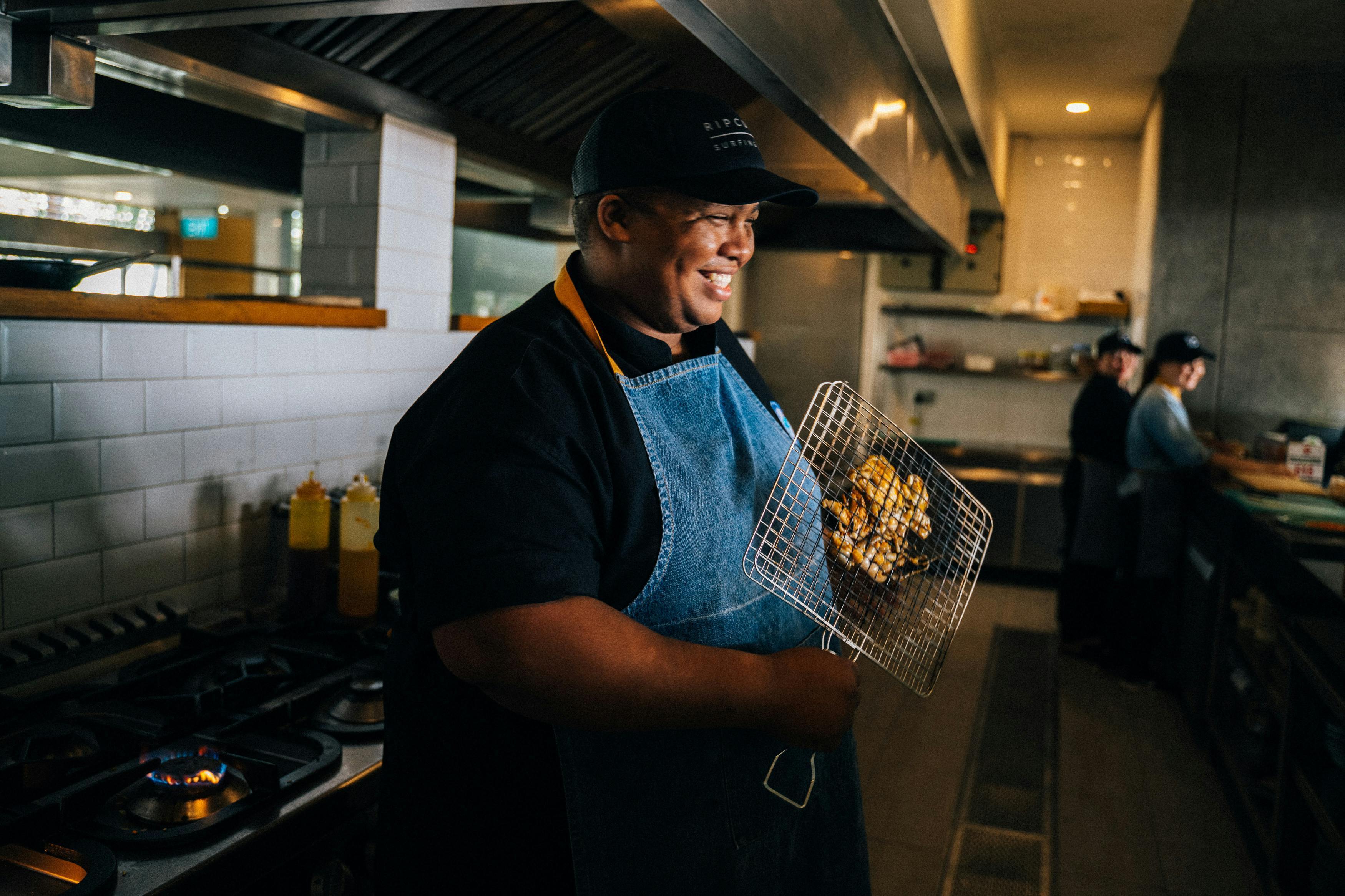 Smiling chef holding a grilling basket of cooked chicken wings in a professional restaurant kitchen, wearing a denim apron and black cap